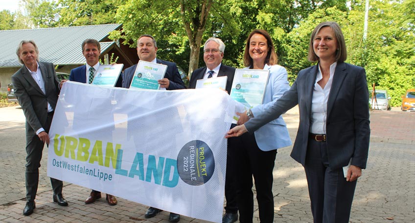 Herbert Weber (Gesch&auml;ftsf&uuml;hrer der OstWestfalenLippe GmbH. L.), &uuml;bergibt die symbolhafte REGIONALE 2022-Auszeichnung in Form von Urkunde und Fahne an die Landr&auml;te Michael Stickeln (Kreis H&ouml;xter), Dr. Axel Lehmann (Kreis Lippe), J&uuml;rgen M&uuml;ller (Kreis Herford) und Landr&auml;tin Anna Katharina B&ouml;lling (Kreis Minden-L&uuml;bbecke) gemeinsam mit Annette Nothnagel (Leiterin der REGIONALE 2022).