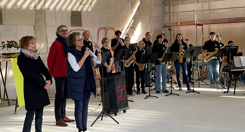 Hoch hinauf musste B&uuml;rgermeister Michael J&auml;cke, um zu pr&uuml;fen, ob der Richtkranz am Rohbau der Sporthalle des Ratsgymnasiums auch richtig h&auml;ngt. Vom rund 8,50 Meter hohen Ger&uuml;st reichte er dem Zimmermann den obligatorischen Schnaps zum Richtspruch. Dieses Ritual verfolgten bei bestem Wetter am 11. November rund 60 G&auml;ste, die die Schule und die Stadt als Bauherrin zum Richtfest eingeladen hatten. Den Auftakt machte in der k&uuml;nftigen Sporthalle die Bigband des Ratsgymnasiums mit dem St&uuml;ck &bdquo;Eye oft he Tiger&ldquo;. Nach den Gru&szlig;worten gaben die Sch&uuml;ler*innen dann noch &bdquo;We are the Champions&ldquo; zum Besten.     &bdquo;Movimens&ldquo; - das ist der Name, den das Ratsgymnasium dem Neubau geben will, wenn er in den Betrieb geht, berichtete Schulleiterin Cordula K&uuml;ppers am vergangenen Freitag. Die Halle befinde sich &bdquo;mitten im Kiez&ldquo; der Altstadt und solle k&uuml;nftig eine offene Sporthalle f&uuml;r das gesamte Quartier werden. Sie w&uuml;nscht sich auch, dass auf dem Kunstrasenplatz, der nach der Inbetriebnahme der Halle erneuert werden muss, wieder viel Fu&szlig;ball gespielt wird. Neben dem Schulsport k&ouml;nne es Angebote wie zum Beispiel Yoga-Kurse geben. Schon vor dem Bau gab es Kooperationen mit der Juxbude, der Volkshochschule, der Eine-Welt-Schule sowie mit anderen Nachbarn, so K&uuml;ppers.  Auf die im August 2021 abgerissene alte Einfach-Sporthalle sollte nicht ein gleich gro&szlig;er Ersatzbau aufgesetzt werden, erl&auml;utert Stadtk&auml;mmerer Norbert Kresse, der auch f&uuml;r die Geb&auml;udewirtschaft verantwortlich ist. Angedockt an die Halle werde es &ndash; nach einem l&auml;ngeren, gemeinsamen Planungsprozess zwischen Stadt und Schule - k&uuml;nftig einen Raum f&uuml;r sporterg&auml;nzende Nutzung wie zum Beispiel Tanzen/Gymnastik/Yoga und f&uuml;r den Sportunterricht auch einen Seminarraum mit Bild- und Filmpr&auml;sentation f&uuml;r den Bereich der Bewegungsanalyse geben. Dieser Raum k&ouml;nne auch von der VHS f&uuml;r beispielsweise Sprachkurse genutzt werden.   Die h&ouml;herwertige Ausstattung der Halle wurde &uuml;ber gesammelte Spenden des F&ouml;rdervereins finanziert. Zahlreiche Mindener Unternehmen und auch Stiftungen h&auml;tten das Projekt gro&szlig;z&uuml;gig unterst&uuml;tzt, wie die Vorsitzende der Gesellschaft der F&ouml;rderer des Ratsgymnasiums e.V., Verena B&ouml;cker, beim Richtfest stolz berichtete.  Der Neubau sei notwendig geworden, weil das Vorg&auml;nger-Modell &bdquo;in die Jahre&ldquo; gekommen war. Die Fassade hatte Risse, was zu einem gro&szlig;en Problem f&uuml;hrte, so B&uuml;rgermeister Michael J&auml;cke in seinem Gru&szlig;wort. Vor dem Hintergrund der vor allem krisenbedingten Mehrkosten bekr&auml;ftigte J&auml;cke vor den G&auml;sten die Entscheidung der Stadt, diese Halle zu bauen. &bdquo;Es ist richtig, dass wir das Projekt auch in schwierigen Zeiten angegangen sind und weiter vorangetrieben haben&ldquo;, so der B&uuml;rgermeister weiter.    Die Arbeiten liegen coronabedingt und aufgrund des andauernden Krieges in der Ukraine, der zu Materialengp&auml;ssen gef&uuml;hrt hat, nicht mehr im Zeitplan. Ferner musste das Baufeld durch den Kampfmittelr&auml;umdienst aufwendig sondiert und untersucht werden. Die deutschlandweit auftretenden Kostenexplosionen am Bau hat auch dieses Projekt - &uuml;ber alle Gewerke hin - hart getroffen: Waren die Planer anfangs noch von Projektkosten in H&ouml;he von 2,6 Millionen Euro ausgegangen, rechnet der Bereichsleiter der st&auml;dtischen Geb&auml;udewirtschaft, J&ouml;rn Schunk, aktuell mit 4,6 Millionen Euro - trotz unver&auml;nderter Planung. Der F&ouml;rderverein der Schule unterst&uuml;tzt den Neubau mit 150.000 Euro.    Bis Ende 2023 soll an der Ecke Rodenbecker Stra&szlig;e/Schwichowwall eine neue Einfachsporthalle mit 1.135 Quadratmetern Nutzfl&auml;che sowie dem angeschlossenen Raum f&uuml;r eine sporterg&auml;nzende Nutzung und den Seminarraum entstehen. Obendrauf kommen ein Gr&uuml;ndach und eine Photovoltaikanlage. Klimafreundlich sind auch die Energiequellen. Geheizt wird die Halle k&uuml;nftig mit einer Luft-Wasser-W&auml;rmepumpe plus Fernw&auml;rme und Solarzellen. &bdquo;Das sei zwar nicht die kosteng&uuml;nstigste Variante, aber &ouml;kologisch und zukunftsweisend&ldquo;, betonte B&uuml;rgermeister J&auml;cke.  Auch architektonisch galt es Herausforderungen zu meistern. So wird die Halle sehr gro&szlig;e Fenster zum Rasenplatz hin haben, um viel nat&uuml;rliches Licht in die Halle zu lassen. Die Fenster korrespondierten zun&auml;chst nicht mit den geforderten Sicherheitsstandards in Sporthallen. Aber auch daf&uuml;r wurde eine L&ouml;sung in Form einer zweiten Glaswand als Prallschutz von den Fachleuten der Geb&auml;udewirtschaft gefunden, so Schulleiterin Cordula K&uuml;ppers. Die Nutzer*innen k&ouml;nnen die Halle &uuml;ber zwei Eing&auml;nge erreichen &ndash; vom S&uuml;den und vom Norden her. Fahrradabstellpl&auml;tze sind im Nordwesten geplant.  Bis die neue Halle fertig ist, m&uuml;ssen die Sportlehrer*innen am Ratsgymnasium weiter kreativ sein. So w&uuml;rden andere Hallen in anderen Stadtbezirken genutzt. &bdquo;Wir fahren jetzt viel Bus&ldquo;, so K&uuml;ppers. Aber es gebe auch Sportunterricht drau&szlig;en, Tischtennis in der Aula und weitere Bewegungs-Ideen. Es lohne sich unbedingt auf die Halle zu warten, meint Cordula K&uuml;ppers. Ihr Dank und auch der des B&uuml;rgermeisters sowie der F&ouml;rdervereinsvorsitzenden galt den Mitarbeitern der st&auml;dtischen Geb&auml;udewirtschaft f&uuml;r die Planungen, Durchf&uuml;hrung und die Projektleitung sowie den &bdquo;flei&szlig;ig arbeitenden&ldquo; Handwerkern.